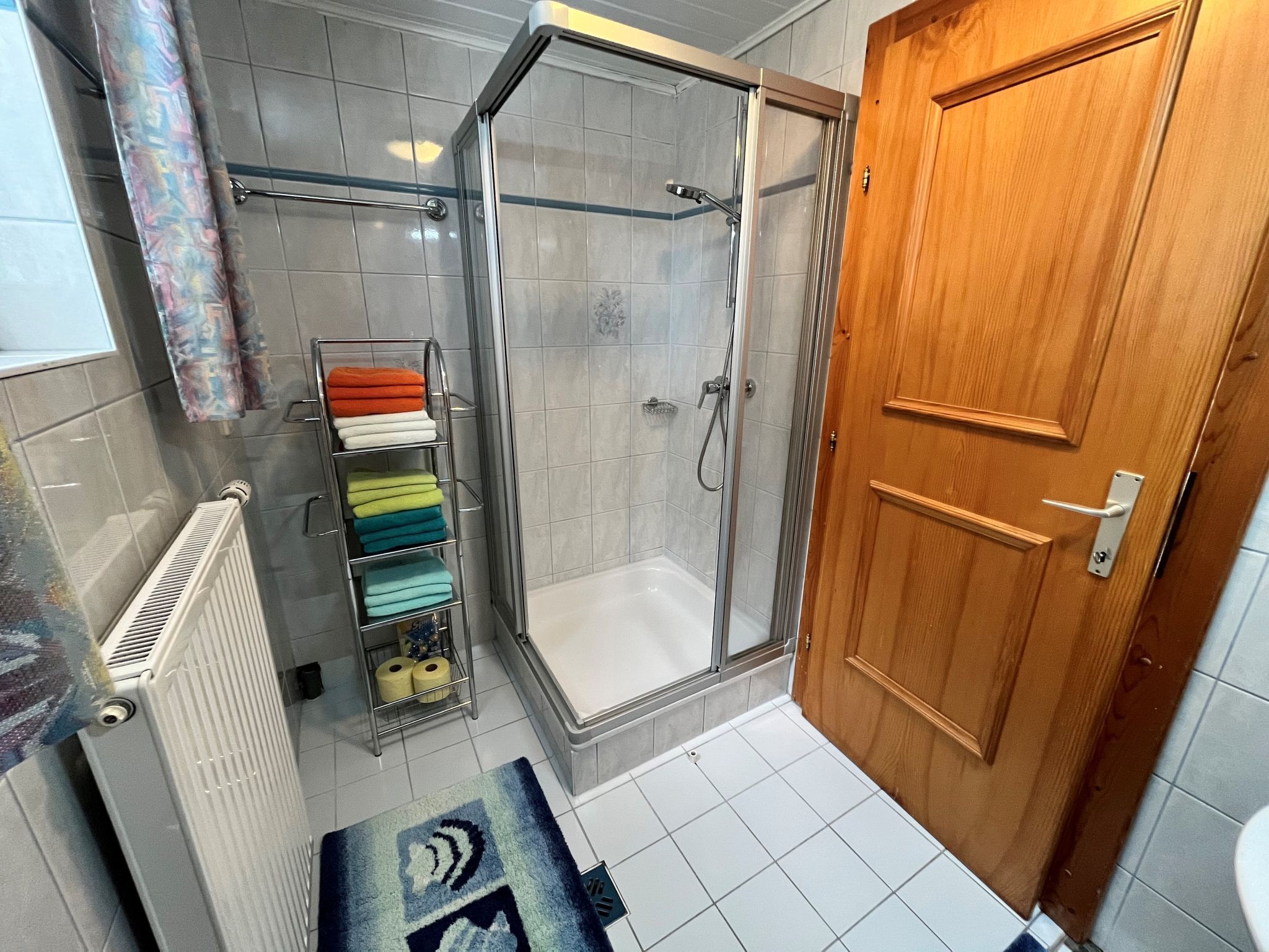 Een badkamer met een douche, een houten deur, een handdoekrek met meerdere handdoeken en een radiator.