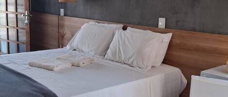 A modern bedroom with a large bed, a wooden headboard, a painting of a coastal scene, and two wall-mounted lamps.