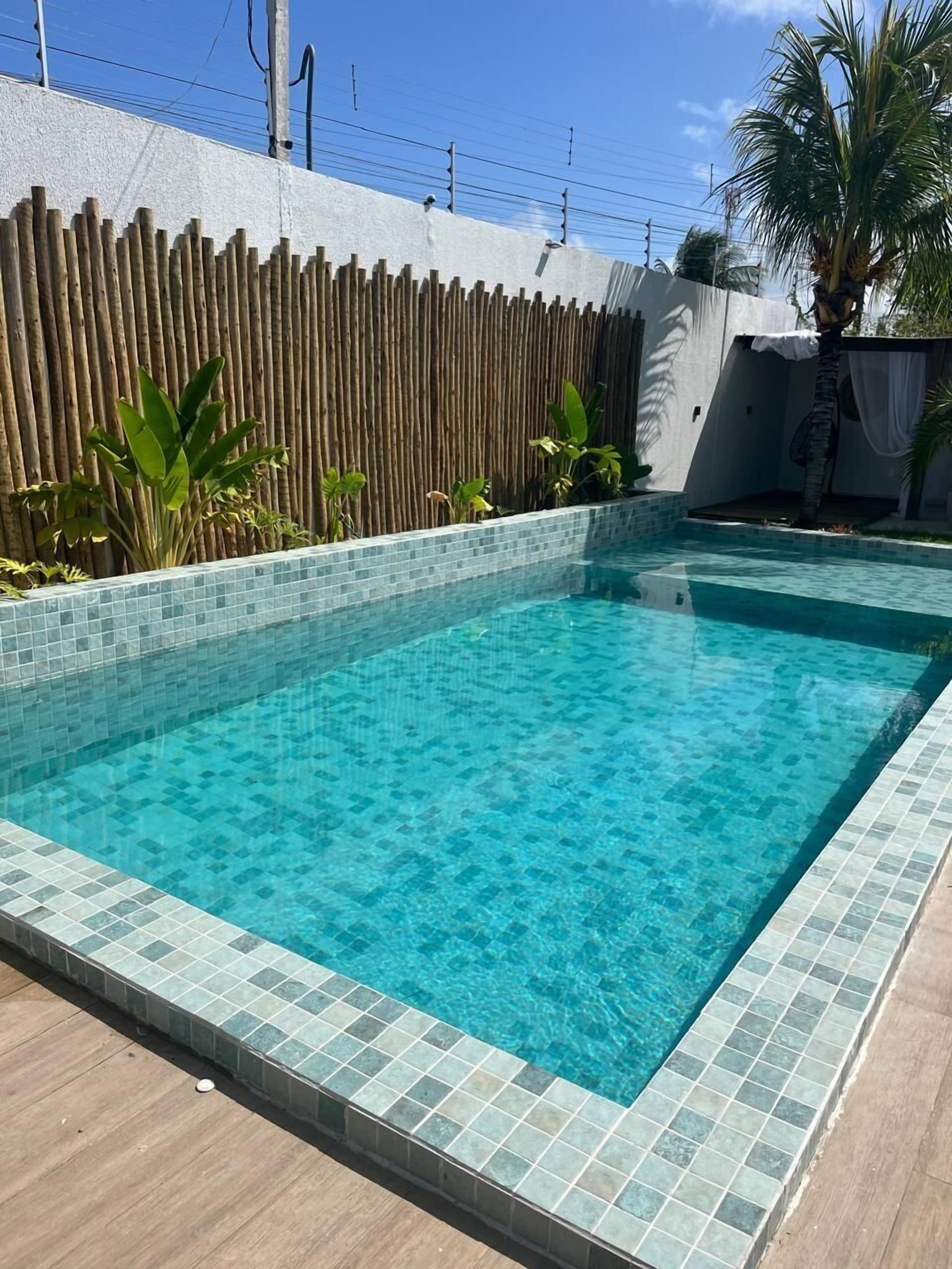 Una piscina rectangular con agua azul cristalina, bordeada por un borde de baldosas y una valla de madera.