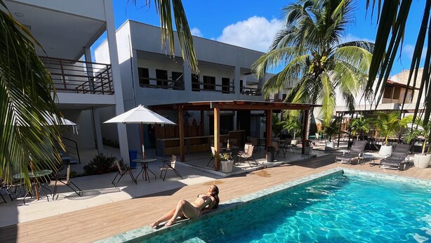 A swimming pool with a person relaxing on the edge, surrounded by lounge chairs and umbrellas, with palm trees and a modern building in the background. - Maré do Frances (Marechal Deodoro)