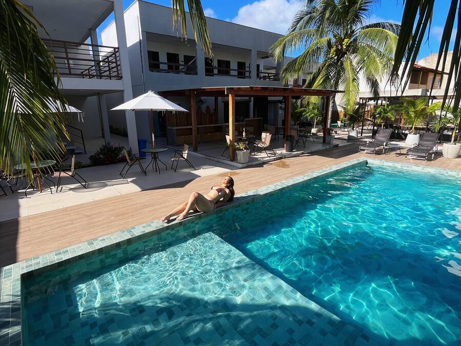 Una piscina con una persona relajándose en el borde, rodeada de sillones de descanso y sombrillas, con palmeras y un edificio moderno al fondo.