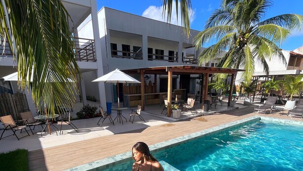 A pool area with lounge chairs and umbrellas, surrounded by palm trees and a modern building. - Maré do Frances (Marechal Deodoro)
