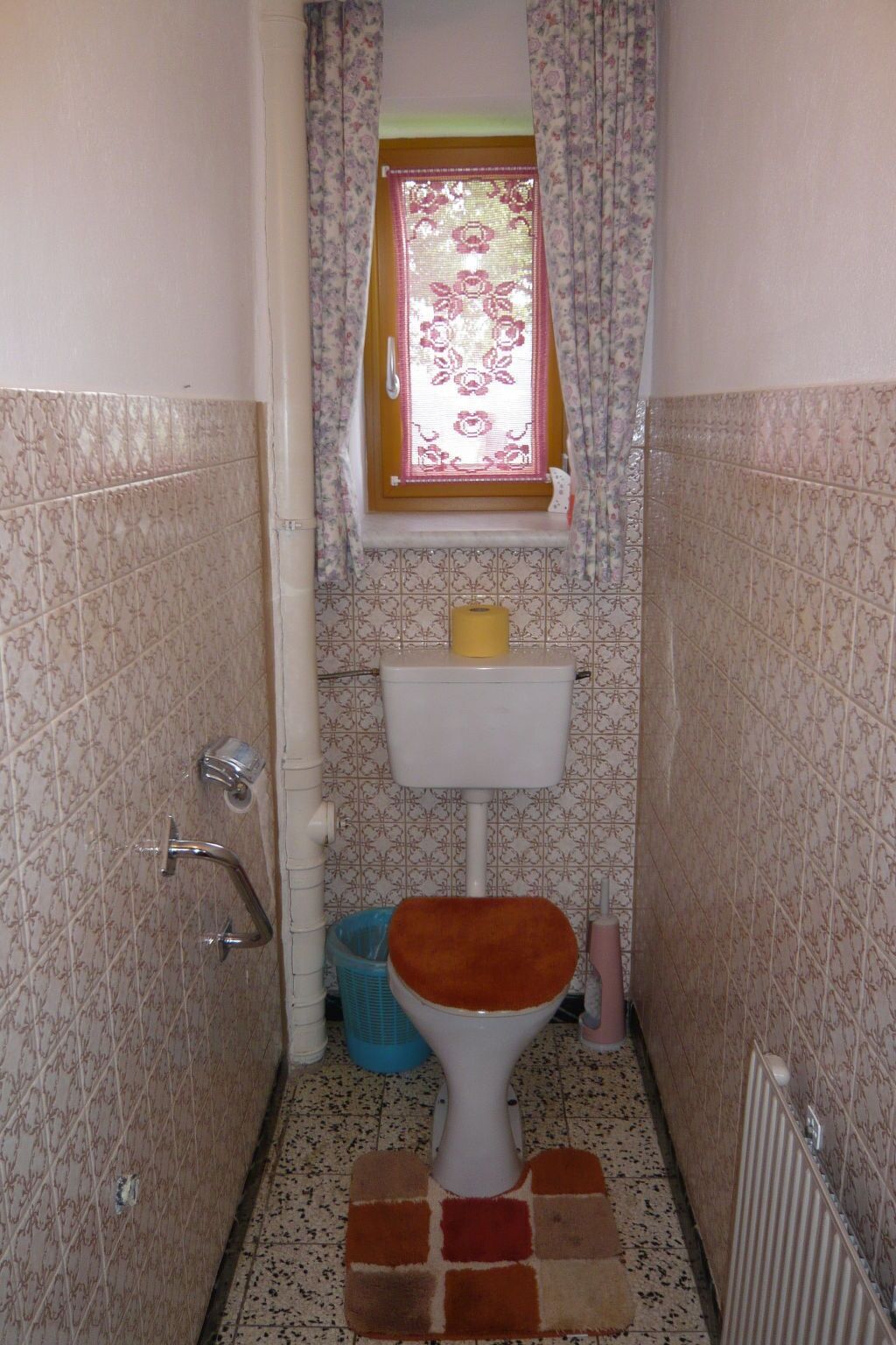A small bathroom with a toilet, a window with floral curtains, and patterned tiles on the walls and floor.