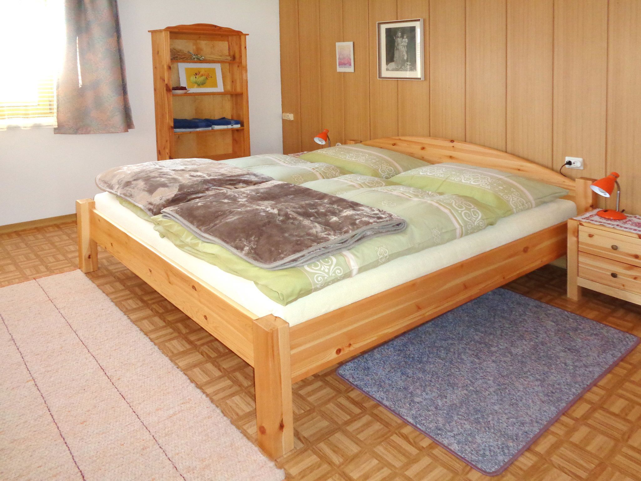 Una cama de madera con colcha verde y blanca, una mesita de noche de madera con lámpara, un armario de madera y un pequeño estante con objetos decorativos.