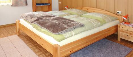 Una cama de madera con colcha verde y blanca, una mesita de noche de madera con lámpara, un armario de madera y un pequeño estante con objetos decorativos.