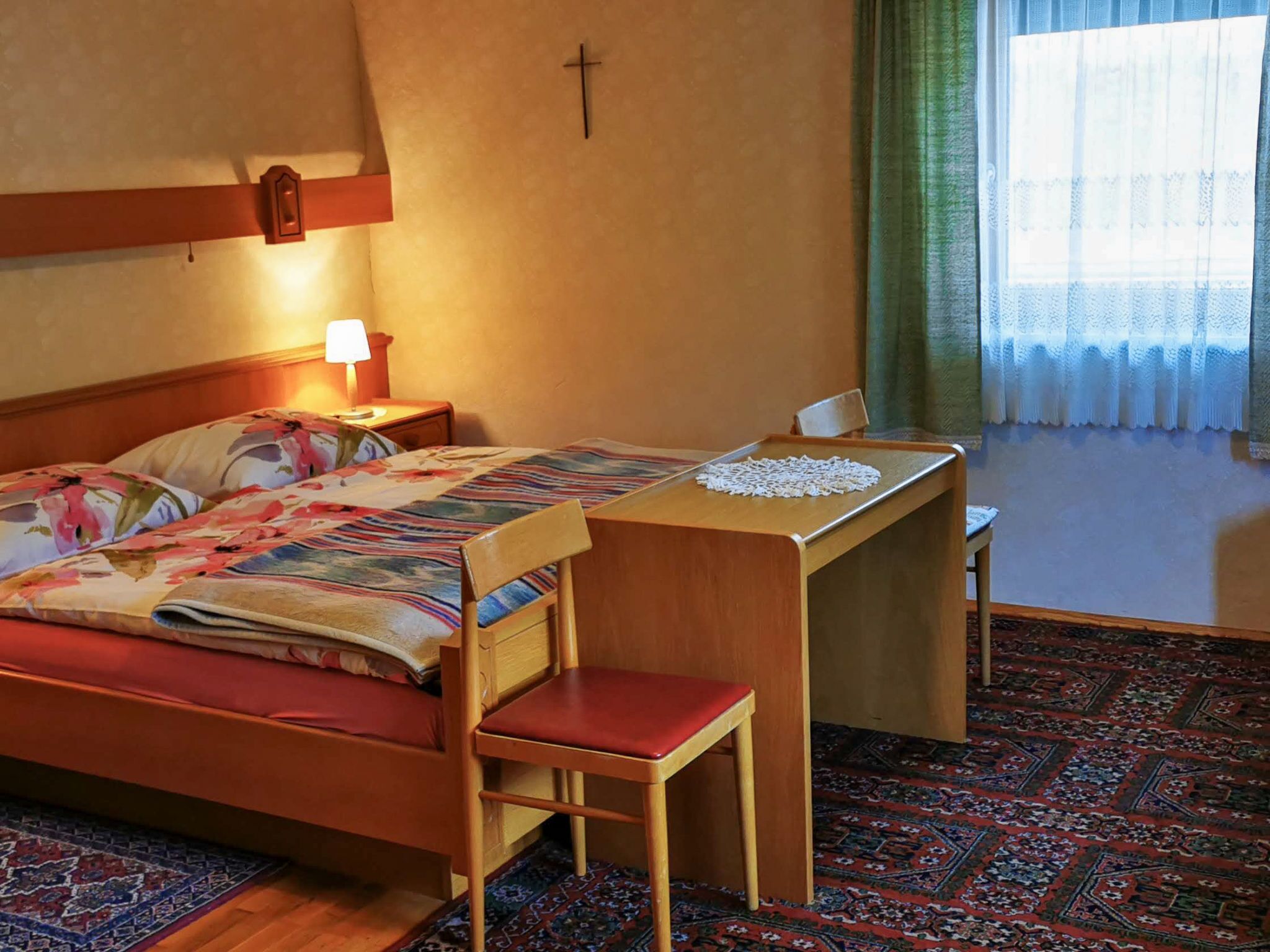 Een hotelkamer met een bed, een bureau, een stoel, een lamp, een kruis aan de muur en een kamerbreed tapijt met een patroon.