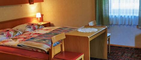 Een hotelkamer met een bed, een bureau, een stoel, een lamp, een kruis aan de muur en een kamerbreed tapijt met een patroon.