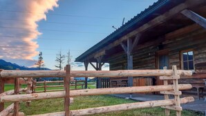 Eine rustikale Holzhütte mit Veranda, umgeben von einem Holzzaun, vor einer Bergkulisse und bewölktem Himmel.
