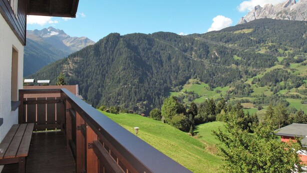 Ein Balkon mit Blick auf eine gebirgige Landschaft, darunter ein bewaldetes Gebiet und ferne Berge.