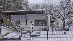 Edificio cubierto de nieve con fachada de madera, una ventana visible y un área cercada con nieve en el suelo.