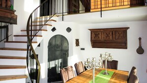 Un comedor con una mesa de madera, sillas y una araña. Se observa una escalera de caracol a la izquierda.