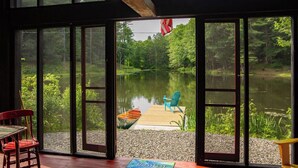 A room with large windows overlooking a lake, a dock with a boat, and outdoor seating. - Catskills Cabin on 100 Wooded Acres with Lake (Climax)