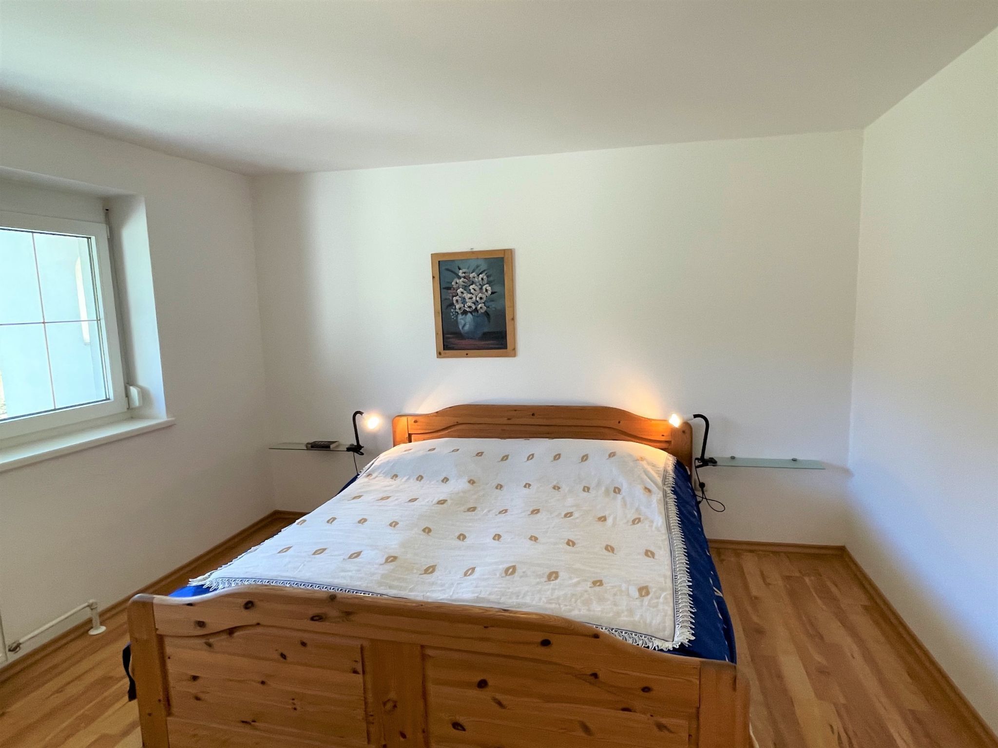 Un lit simple avec un cadre en bois et une couvre-lit blanche, dans une pièce aux murs blancs et au plancher en bois.