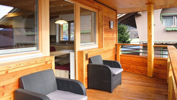 Una terraza de madera con dos sillas de mimbre y una mesa, ventanas amplias y vistas al exterior.