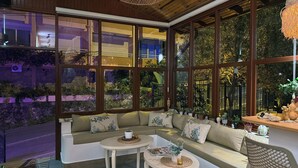 Un salon moderne avec une grande fenêtre, un canapé, deux petites tables et un fauteuil en osier.