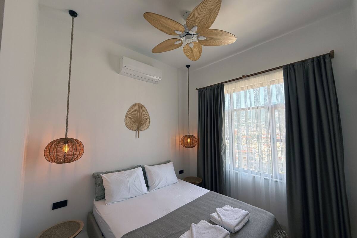 Une chambre d’hôtel comprenant un lit, deux tables de chevet, un ventilateur de plafond, un climatiseur et une fenêtre avec des rideaux.