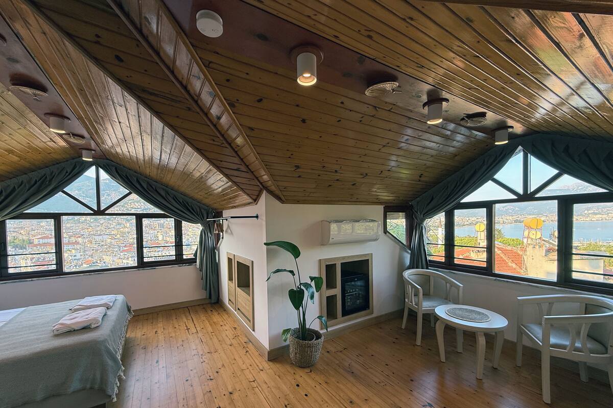 Une pièce avec un plancher en bois, un lit, une table à manger avec des chaises, une plante en pot et de grandes fenêtres donnant sur la ville.