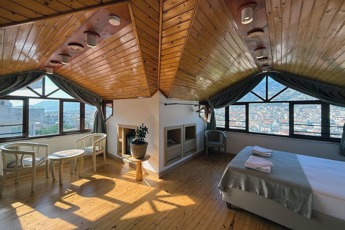 Une pièce avec des poutres apparentes en bois au plafond, un lit, une petite table avec une plante, une chaise et une vue sur la ville grâce à de grandes fenêtres.