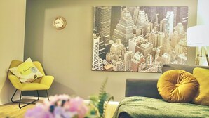 Um quarto com uma pintura de paisagem urbana, um relógio, uma cadeira amarela e um sofá verde.