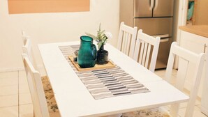 Uma área de refeições com uma mesa branca, cadeiras estampadas com flores, um jarro verde e uma estante com plantas.