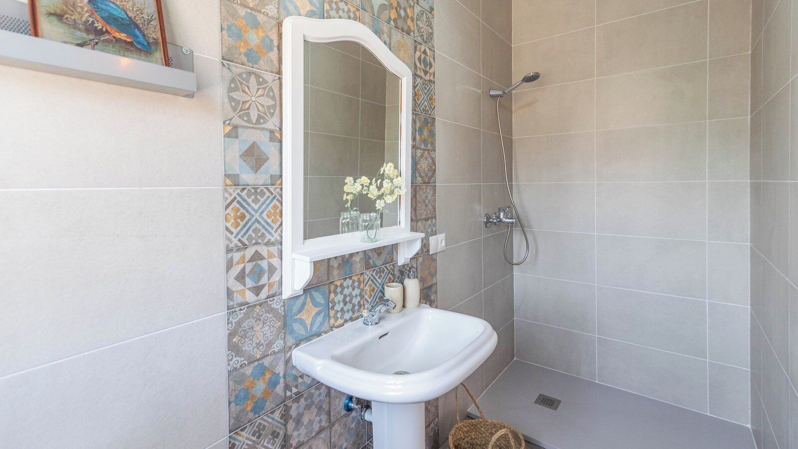 Um banheiro com pia branca, espelho e chuveiro. As paredes têm azulejos estampados.