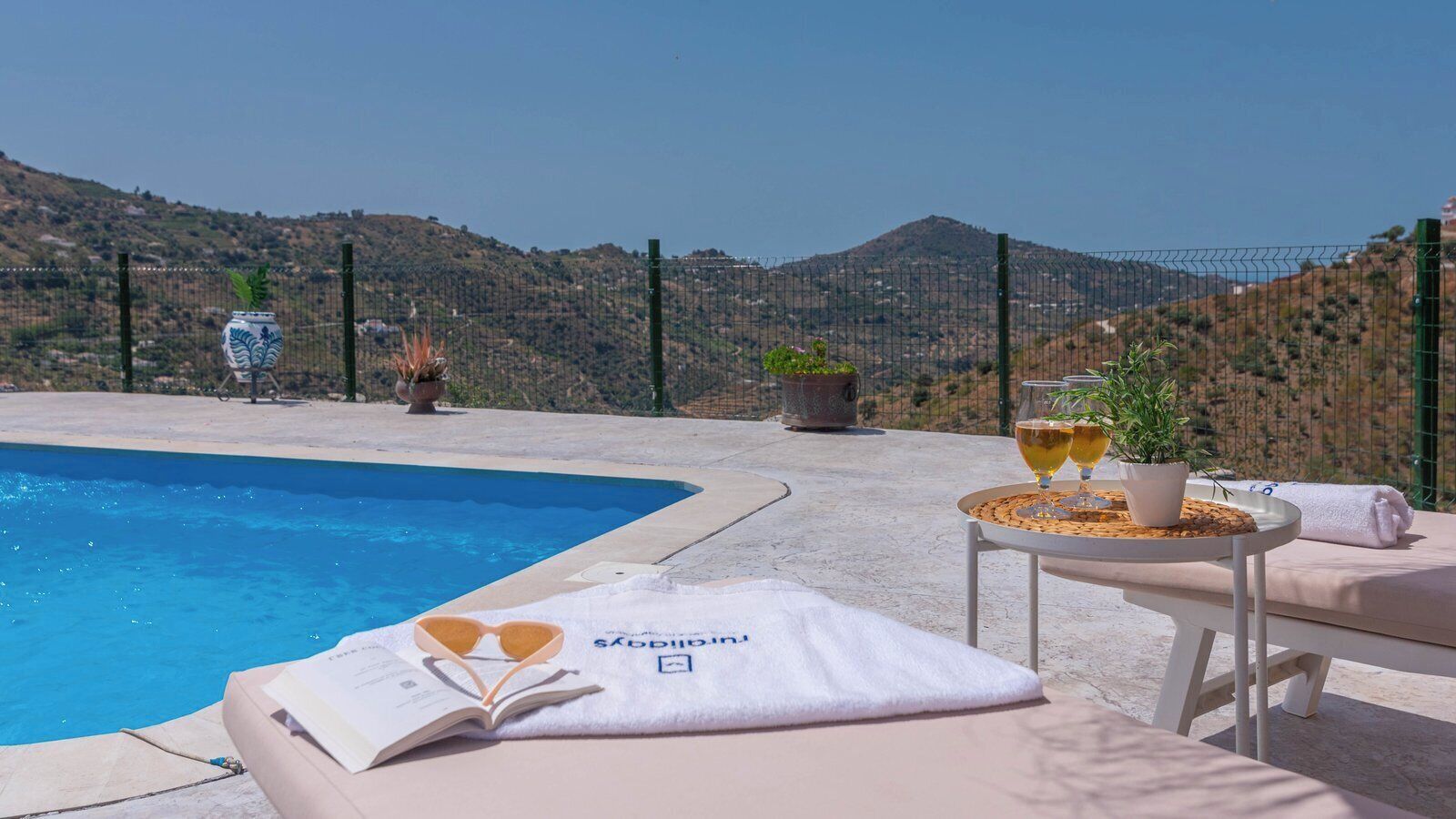 A poolside area with lounge chairs, a table with drinks, and a view of the hills.