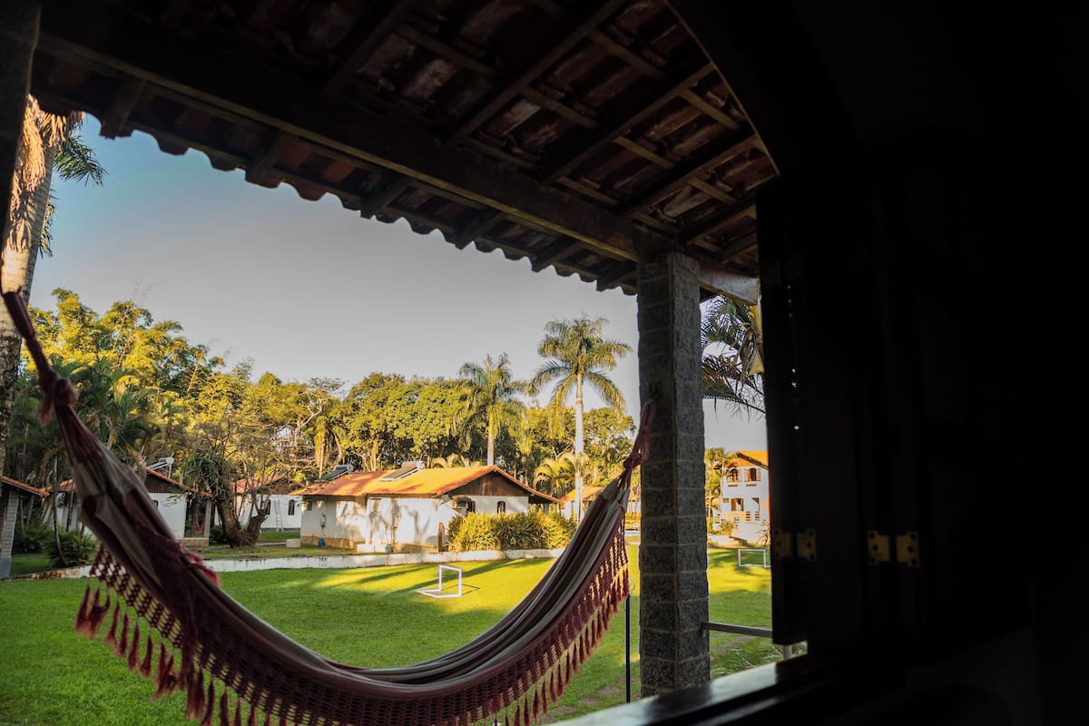 A hammock under a wooden structure with a view of a well-maintained lawn, palm trees, and buildings in the background.