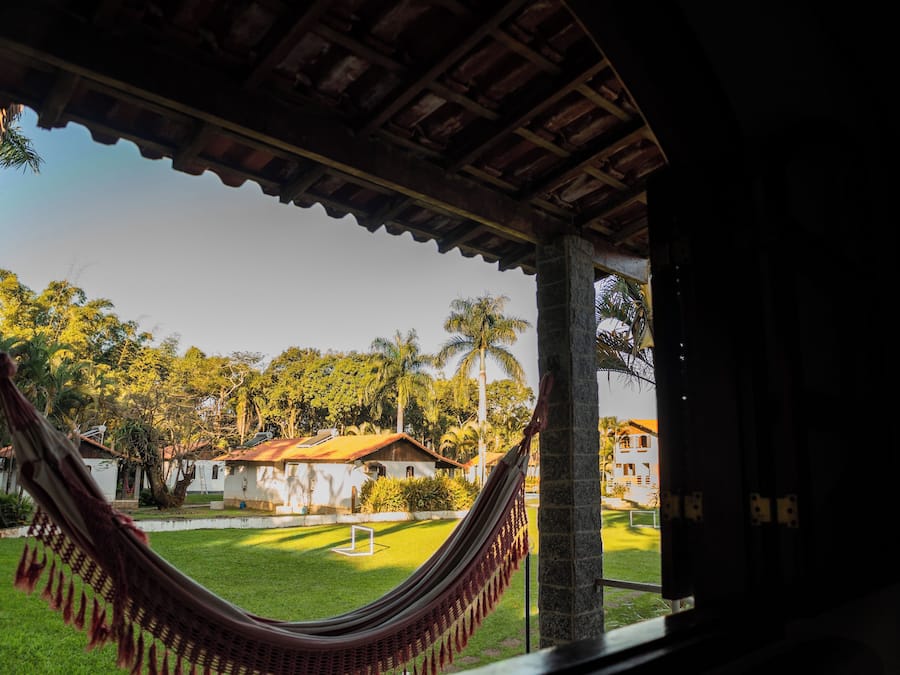 Una hamaca bajo una estructura de madera con vista a un césped cuidado, palmeras y edificios al fondo.