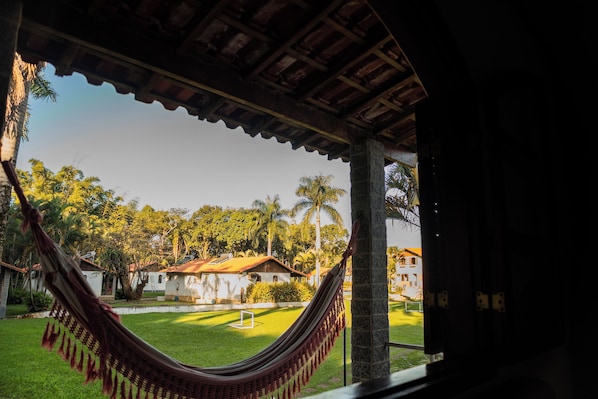 Una hamaca bajo una estructura de madera con vista a un césped cuidado, palmeras y edificios al fondo.