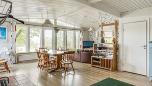 A spacious living area with a wooden dining table, chairs, and a sofa. There is a television, a bookshelf, and a large window providing natural light.