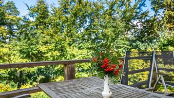Une table en bois sur laquelle est posé un vase blanc contenant des baies rouges et des feuilles vertes. La scène se déroule à l’extérieur, avec une clôture en bois et des arbres en arrière-plan.