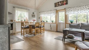 Een ruime woonkamer met een houten eettafel, stoelen, een bank en een bijzettafeltje. Er zijn grote ramen met bloemen gordijnen, een plafondventilator en een schilderij aan de muur.