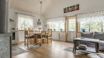 Un vaste espace de vie comprenant une table à manger en bois, des chaises, un canapé et une table basse. De grandes fenêtres sont ornées de rideaux à motifs floraux, et on y trouve également un ventilateur de plafond et un tableau accroché au mur.