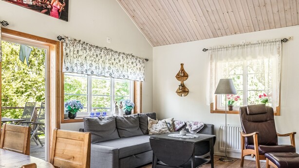 Een gezellige woonkamer met een grijze bank, een houten eettafel en een zwarte salontafel. Er zijn twee stoelen, een lamp en een raam met gordijnen.