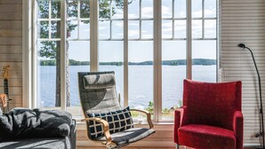 Una habitación con un gran ventanal con vistas a un lago, que incluye una silla roja, un sillón gris y una lámpara de pie negra.