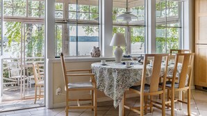 Un comedor con una mesa dispuesta para cuatro personas, rodeada de sillas de madera, ventanas amplias con vistas a árboles y agua, y un ventilador de techo.