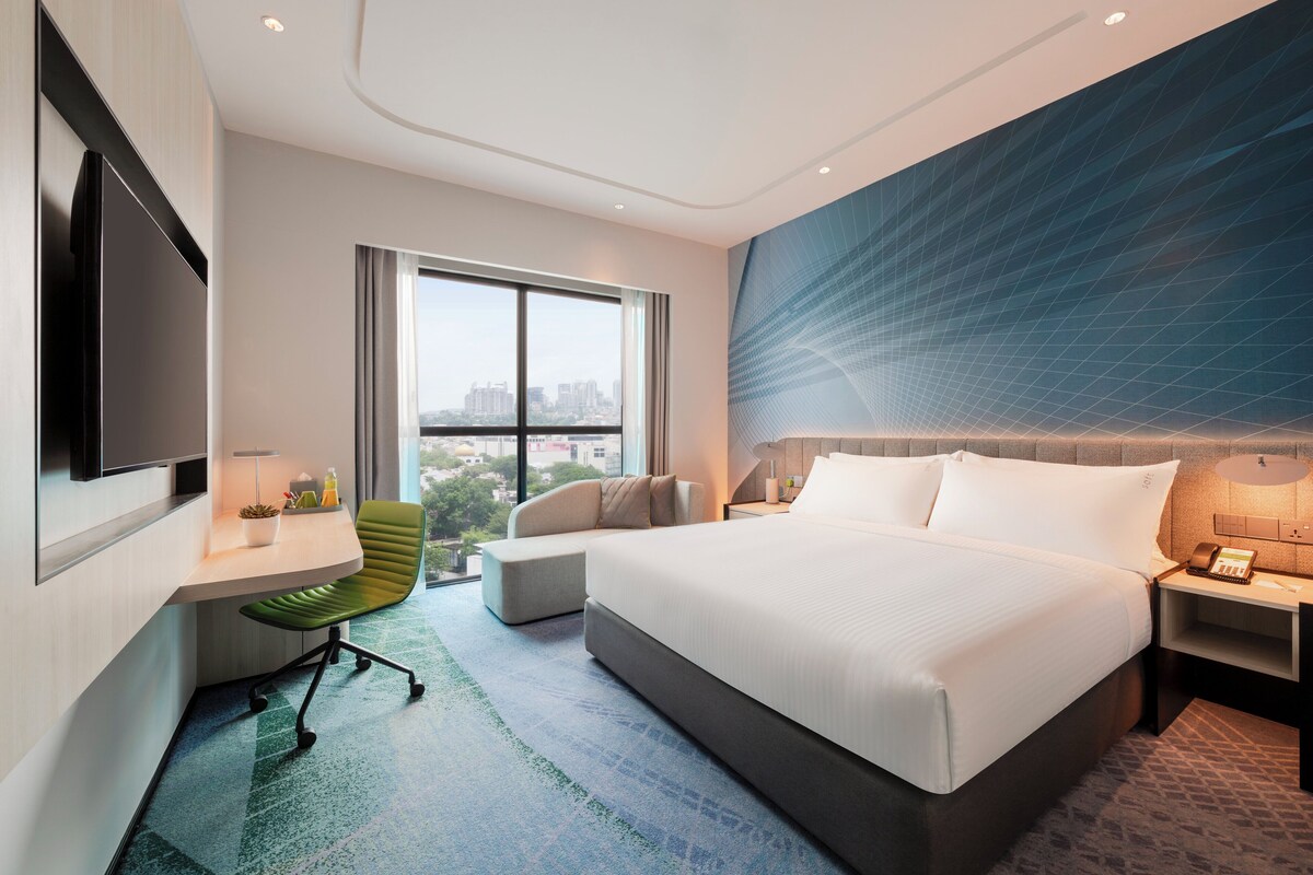 A modern hotel room with a large bed, a desk with a chair, a flat-screen TV, and a large window offering a city view.