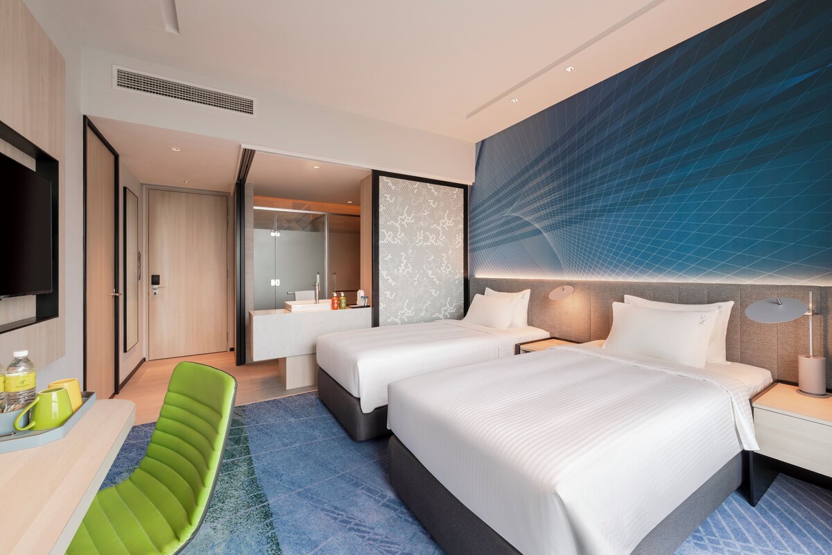 A modern hotel room with two beds, a desk, a green chair, and a large wall mural.