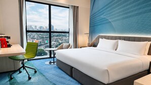 A hotel room with a large bed, a desk, a green chair, and a view of a cityscape through large windows. - Holiday Inn Kuala Lumpur Bangsar by IHG (Kuala Lumpur)