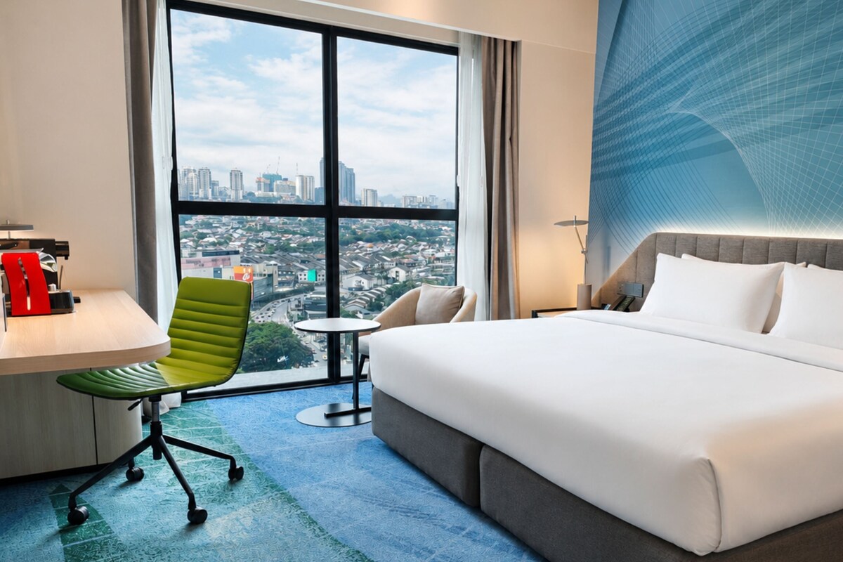 A hotel room with a large bed, a desk, a green chair, and a view of a cityscape through large windows.