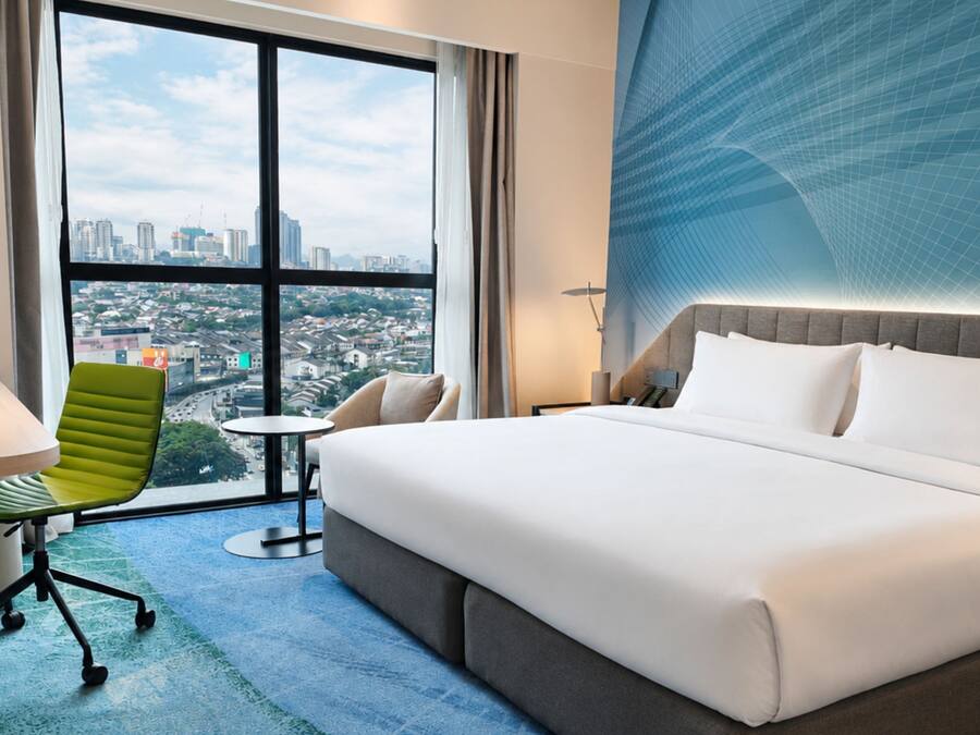 A hotel room with a large bed, a desk, a green chair, and a view of a cityscape through large windows.