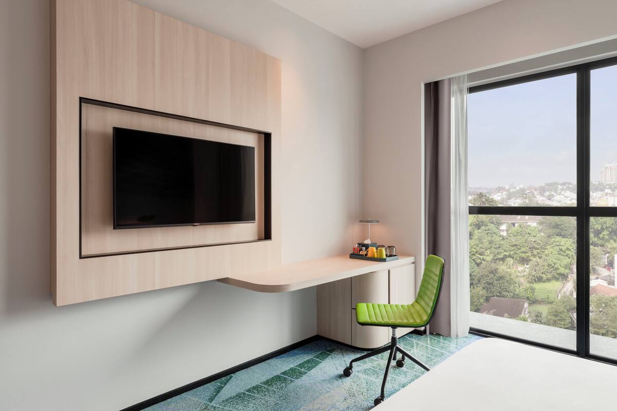 A modern hotel room with a mounted TV, a desk with a lamp, a green chair, and a large window offering a city view.