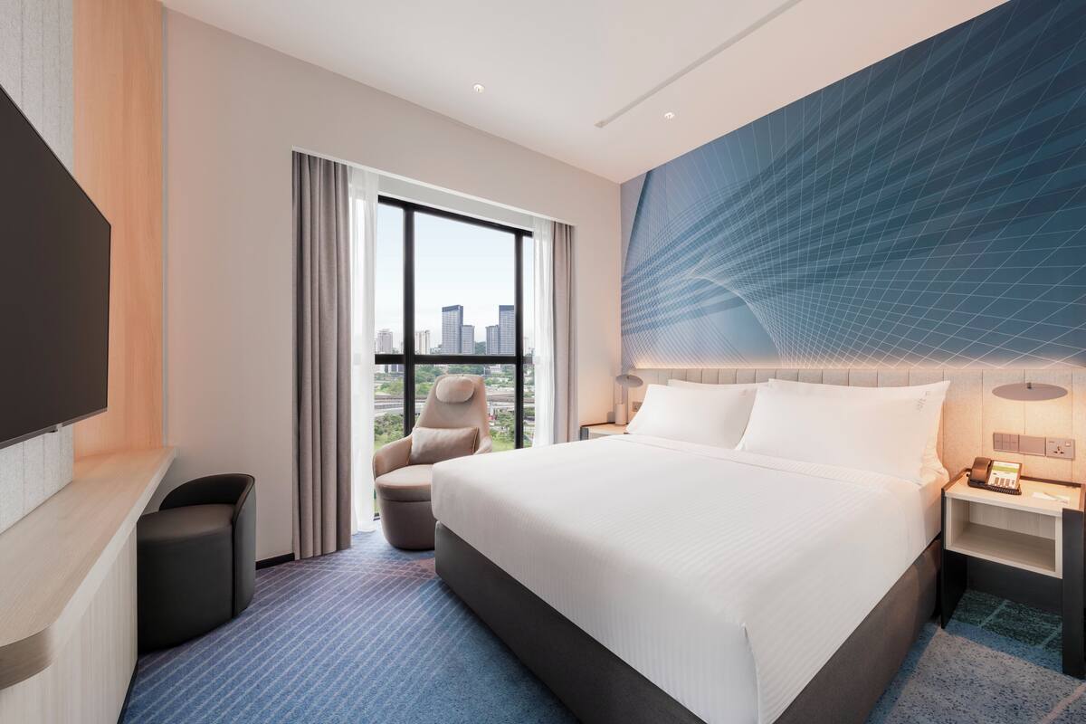 A hotel room with a large bed, a desk, a chair, a TV, and a city view.