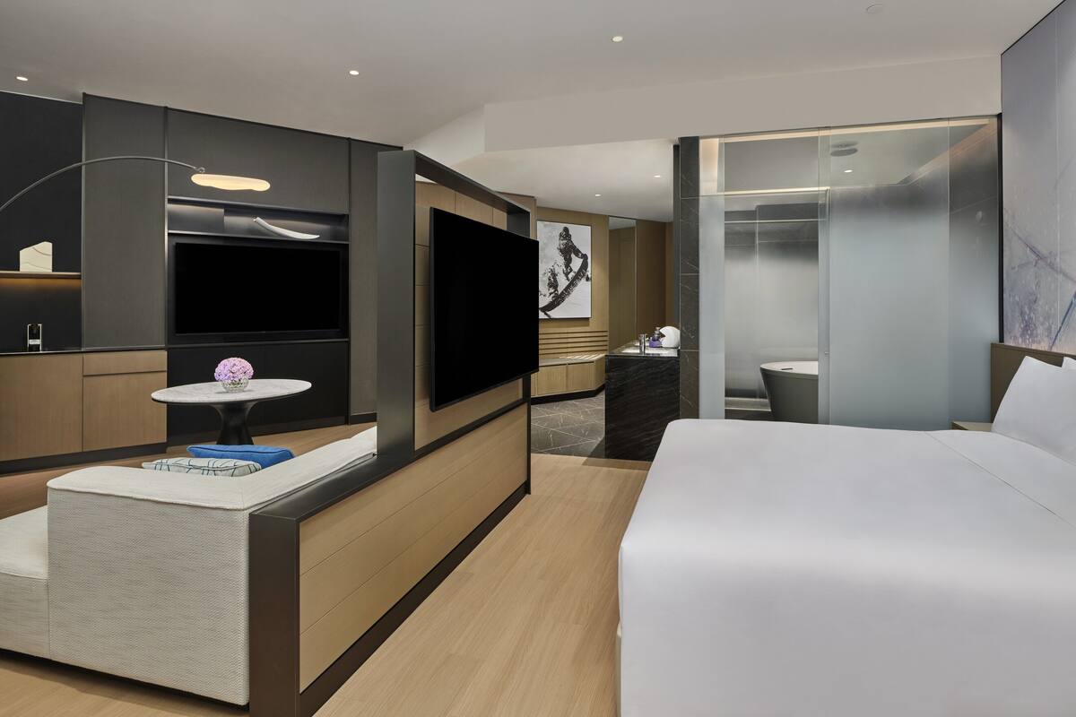 A modern hotel room with a large bed, a flat-screen TV, a small round table with a vase, and a seating area with a sofa and cushions.
