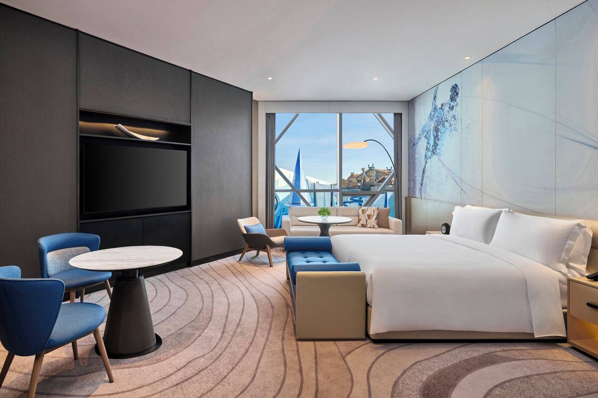 A modern hotel room with a large bed, a seating area with chairs and a table, a TV mounted on the wall, and a large window with a view of a bridge and buildings.