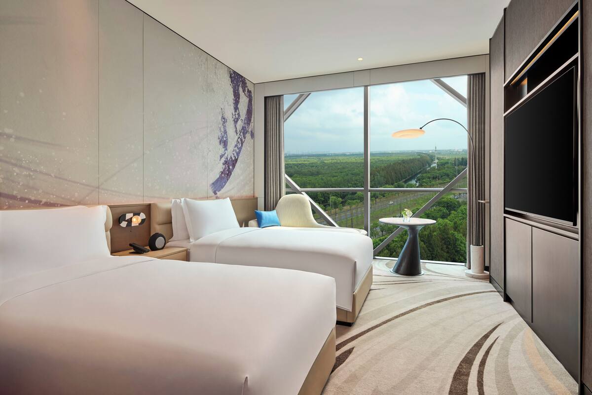 A modern hotel room with two beds, a large window with a city view, a small table, and a TV.
