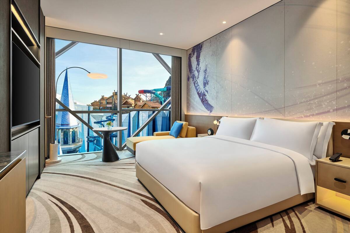 A modern hotel room with a large bed, a small table, and a view of a water park.