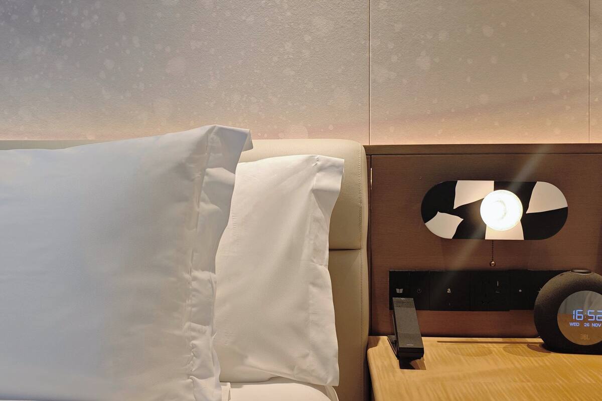 A hotel room with a bed, a nightstand with a lamp, and a wall clock displaying the time.