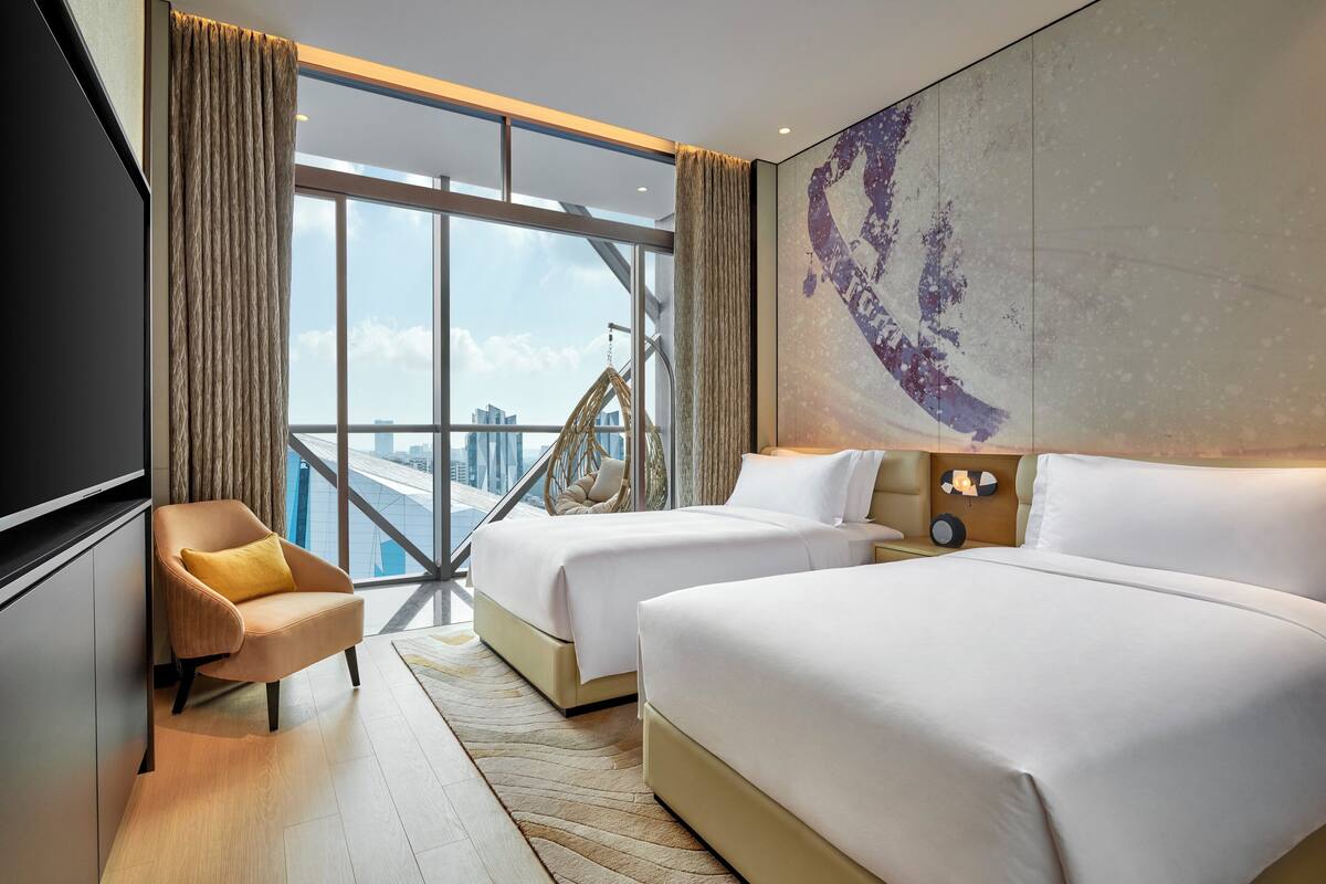 A hotel room with two beds, a sofa, a large window with a view of a cityscape, and a mounted artwork on the wall.