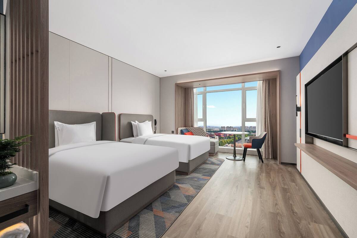 Ein Hotelzimmer mit zwei Betten, einem Fernseher, einem großen Fenster mit Stadtblick und einem kleinen Balkon.